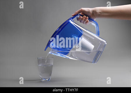 Eine Hand gießen gefiltertes Wasser in das Glas, auf grauem Hintergrund Stockfoto