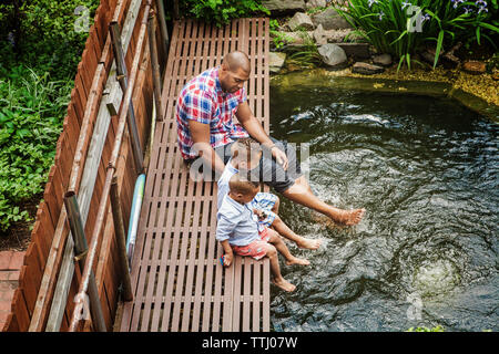 Hohe Betrachtungswinkel von Vater mit Söhnen sitzt auf Fußweg über Teich im Hinterhof Stockfoto