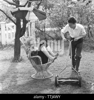 Sommer Aktivität in den 1950er Jahren. Eine junge Frau sitzt im typischen 50er Jahre garten Stuhl ihre Füße oben hält, so dass Ihr Partner das Gras schneiden kann, wo sie sitzt. Schweden Mai 1959 Stockfoto