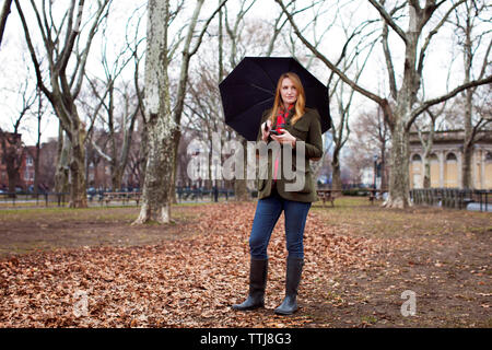 Frau mit Regenschirm weg schauen, während Sie Handy im Park Stockfoto