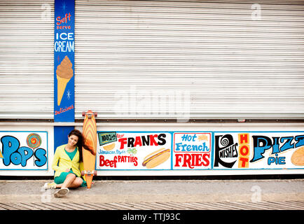 Portrait von jugendlichen Mädchen mit Skateboard gegen geschlossene Food Stores sitzen Stockfoto