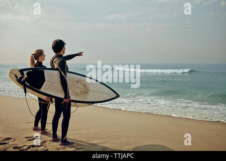 Paar mit Blick auf Meer, während Surfboard am Strand tragen Stockfoto