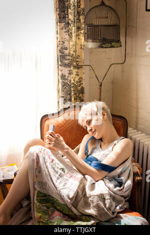 Frau mit Smart Phone während auf Stuhl zu Hause sitzen Stockfoto