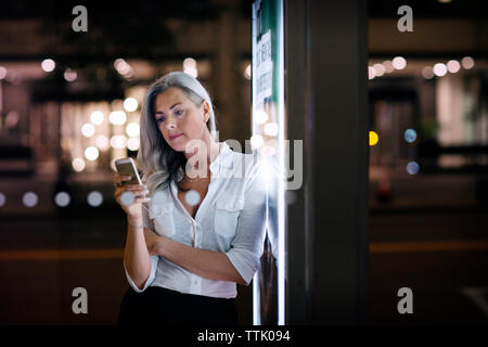 Geschäftsfrau, die ihr Telefon benutzt, während sie sich in der Stadt auf das Poster der Bushaltestelle lehnt Stockfoto