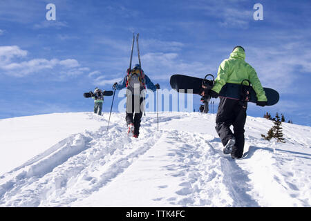 Rückansicht der Freunde, Snowboards und Skier beim Gehen auf Schnee bedeckten Berg Stockfoto