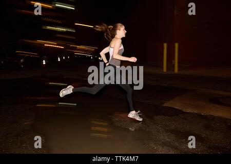 Seitenansicht der weiblichen Jogger springen über Pfütze auf der Straße in der Stadt bei Nacht Stockfoto