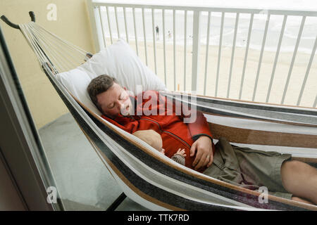 Vater und Sohn ruht in der Hängematte auf dem Balkon, durch das Fenster gesehen Stockfoto