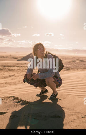 Frau mit Rucksack hockend auf Sand am Strand gegen Himmel bei Sonnenuntergang Stockfoto