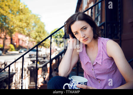 Portrait von Frau mit Kaffeetasse sitzend auf Schritte außerhalb des Gebäudes Stockfoto