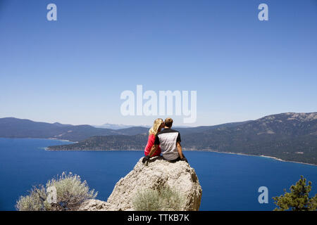 Ansicht der Rückseite des Paar sitzt auf einer Klippe mit Blick auf den See gegen den klaren blauen Himmel Stockfoto