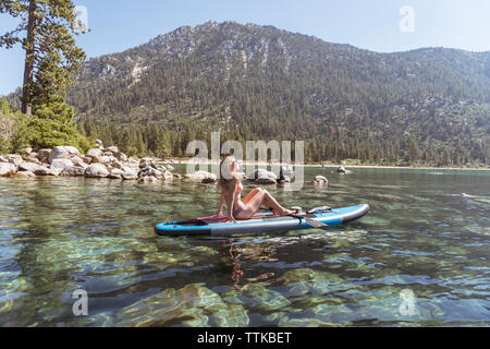 Seitenansicht der Frau im Bikini paddleboarding auf See gegen Berg während der sonnigen Tag Stockfoto