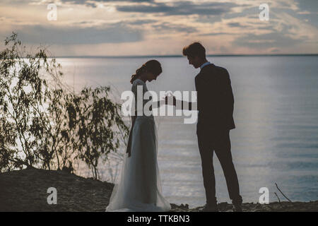 Hochzeitspaar hält Hände, während er am Strand gegen den Himmel während Sonnenuntergang steht Stockfoto