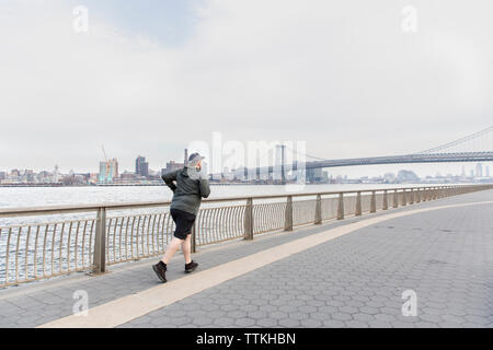 Die volle Länge der übergewichtigen Mann joggen am Fluss mit Williamsburg Bridge im Hintergrund Stockfoto