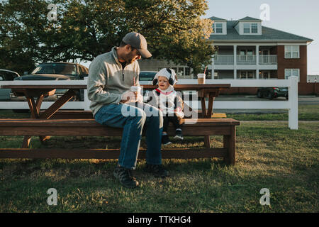 Vater holding Kaffeetasse, während mit Sohn auf Holzbank im Hof sitzen Stockfoto