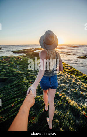 7/8 Hand hand Mann mit Freundin beim Gehen auf Gras am Strand bei Sonnenuntergang Stockfoto