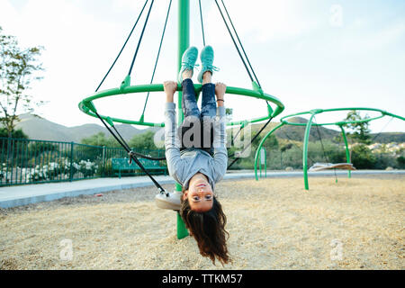 Portrait von Mädchen hing kopfüber an outdoor Spielgeräte gegen Himmel am Spielplatz Stockfoto