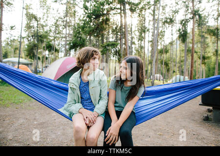 Die Schwestern sprechen beim Sitzen auf der Hängematte gegen Bäume im Wald Stockfoto