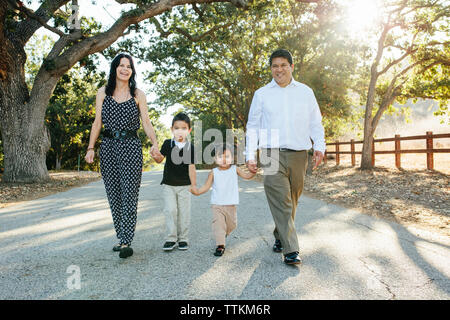 Portrait von Happy Family, Hände halten, beim Gehen auf die Straße gegen Bäume im Park Stockfoto