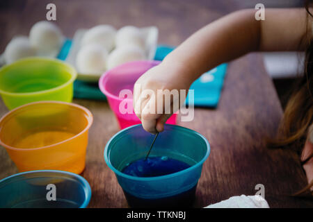 Abgeschnittene Hand eines Mädchens mit Rührer, um Farbe in Wasser für ostereier Färbung auf dem Tisch zu mischen Stockfoto