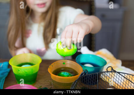 Mitte des Mädchens, das osterei in gefärbtes Wasser taucht, auf dem Tisch zu Hause Stockfoto