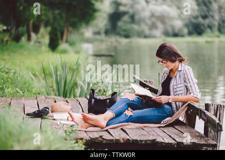 Frau mit Buch beim Sitzen auf Pier gegen See im Wald Stockfoto