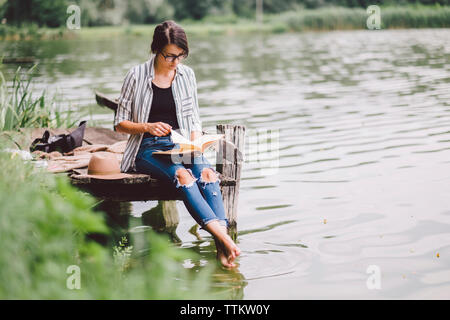 Frau mit Buch beim Sitzen auf Pier über den See im Wald Stockfoto