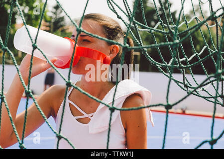 Junge Frau trinkt Wasser aus der Flasche am Fußballplatz durch Netz gesehen Stockfoto