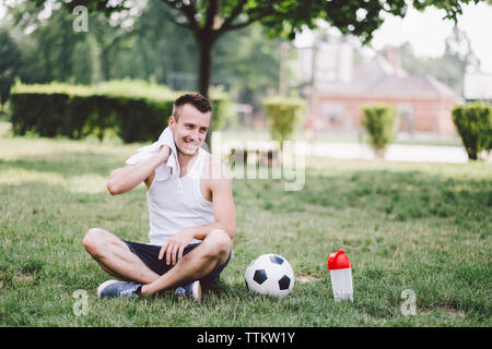 Lächelnden jungen Mann wischen Hals sitzend auf Fußball im Park Stockfoto