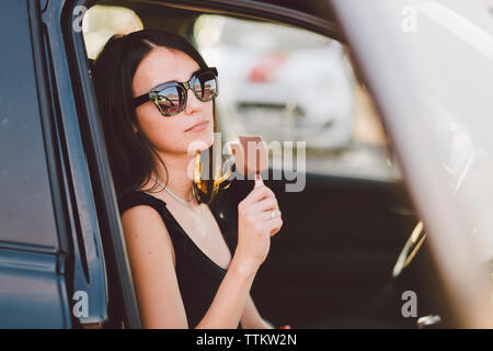 Junge Frau mit Sonnenbrille Holding gefroren süße Speisen beim Sitzen im Auto durch das Fenster gesehen Stockfoto