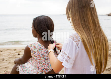 Ansicht der Rückseite Frau Flechten des Freundes Haar, während am Ufer am Strand sitzen Stockfoto