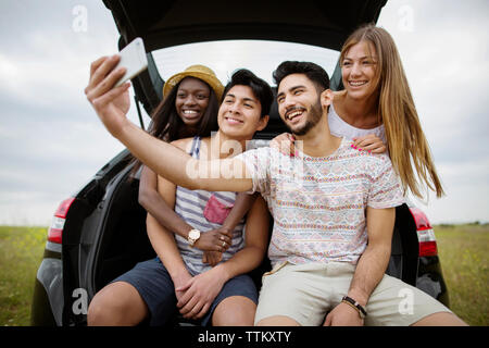 Glücklich, multi-ethnischen Freunde unter selfie durch Smart Phone während der Sitzung in Kofferraum auf dem Feld Stockfoto