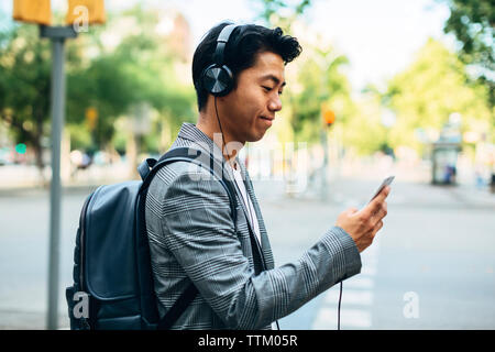 Geschäftsmann mit Rucksack mit Smartphone, während er Musik auf dem Fußweg hört Stockfoto