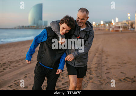 Verspielter Vater mit Arm um die Schulter des Sohnes, der gegen das Hotel Vela am Strand läuft Stockfoto