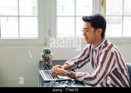 Geschäftsmann, der am Laptop arbeitet, während er im Büro vor den Fenstern sitzt Stockfoto