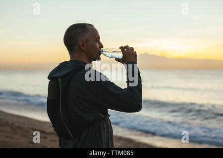 Seitenansicht des Menschen Trinkwasser beim Stehen am Strand bei Sonnenuntergang Stockfoto