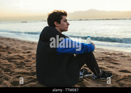 Seitenansicht von nachdenklich Teenager holding Wasserflasche beim Sitzen am Strand Stockfoto