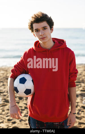 Portrait von zuversichtlich Teenager halten Fußball beim Stehen am Strand gegen Meer Stockfoto