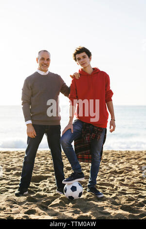 Portrait von zuversichtlich, Sohn und Vater mit Fußball am Strand gegen den klaren Himmel Stockfoto