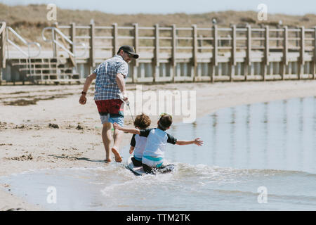 Rückansicht der glückliche Vater ziehen Kinder sitzen auf Surfbrett im Meer während der sonnigen Tag Stockfoto