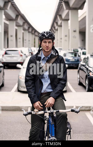Portrait von zuversichtlich Geschäftsmann sitzt auf Fahrrad mit Brücken und Parkplatz im Hintergrund Stockfoto