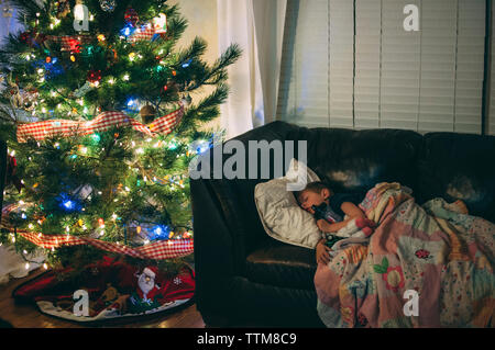 Junge, der nachts auf dem Sofa am beleuchteten weihnachtsbaum schläft Stockfoto