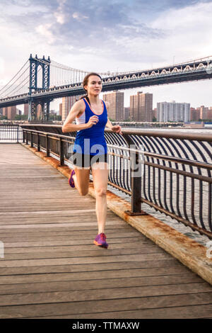 Gesunde junge Frau läuft auf der Brooklyn Bridge Park Promenade mit den East River und die Brooklyn Bridge im Hintergrund, Stockfoto