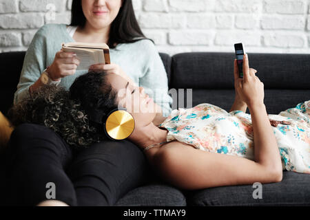 Frau mit Handy beim Entspannen auf dem Schoß der Freundin lesen Buchen Sie im Wohnzimmer Stockfoto