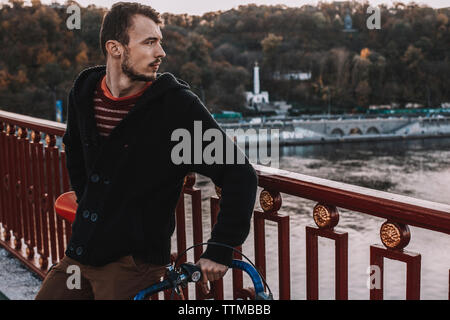 Nachdenklicher Mann weg suchen, während man mit dem Fahrrad auf Brücke über Fluss gegen Bäume bei Sonnenuntergang Stockfoto