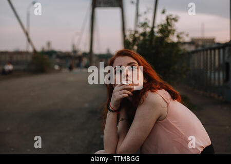 Junges Mädchen mit Sommersprossen rauchen Zigarette beim Sitzen auf der Brücke Stockfoto