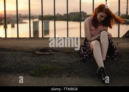 Junge rothaarige Frau mit Smart Phone, während auf der Brücke in der Stadt sitzen Stockfoto