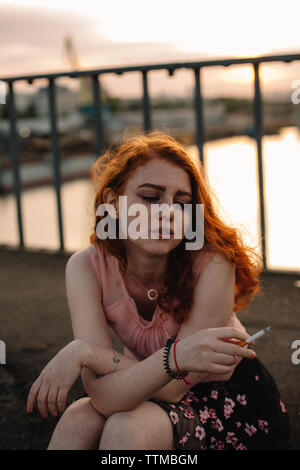 Junge rothaarige Frau rauchen Zigarette beim Sitzen auf der Brücke in der Stadt Stockfoto