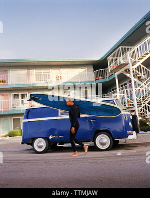 USA, Kalifornien, Capitola, ein Surfer mit einem blauen longboard Spaziergänge in fronr eines blauen VW-Bus auf dem Weg zum Surfen Stockfoto