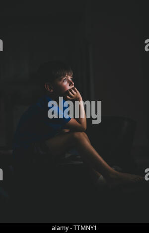 Seitenansicht der nachdenkliche Junge mit der Hand am Kinn sitzen auf Stuhl in der Dunkelkammer Stockfoto