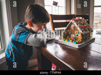 Glücklicher Junge, der während weihnachten auf einem Holztisch Lebkuchenhaus schaut Stockfoto